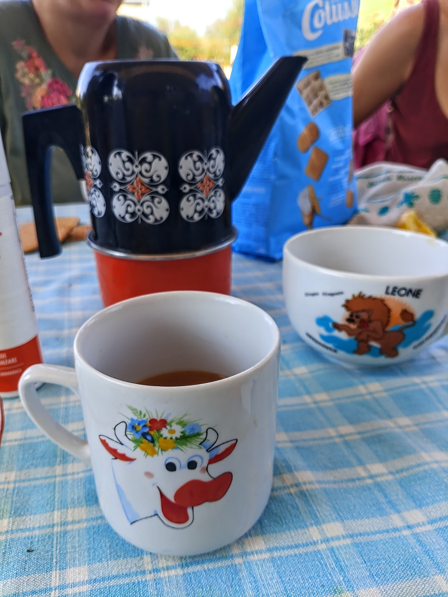 A Kinder Lada cow mug beside a coffee pot on a checked tablecloth