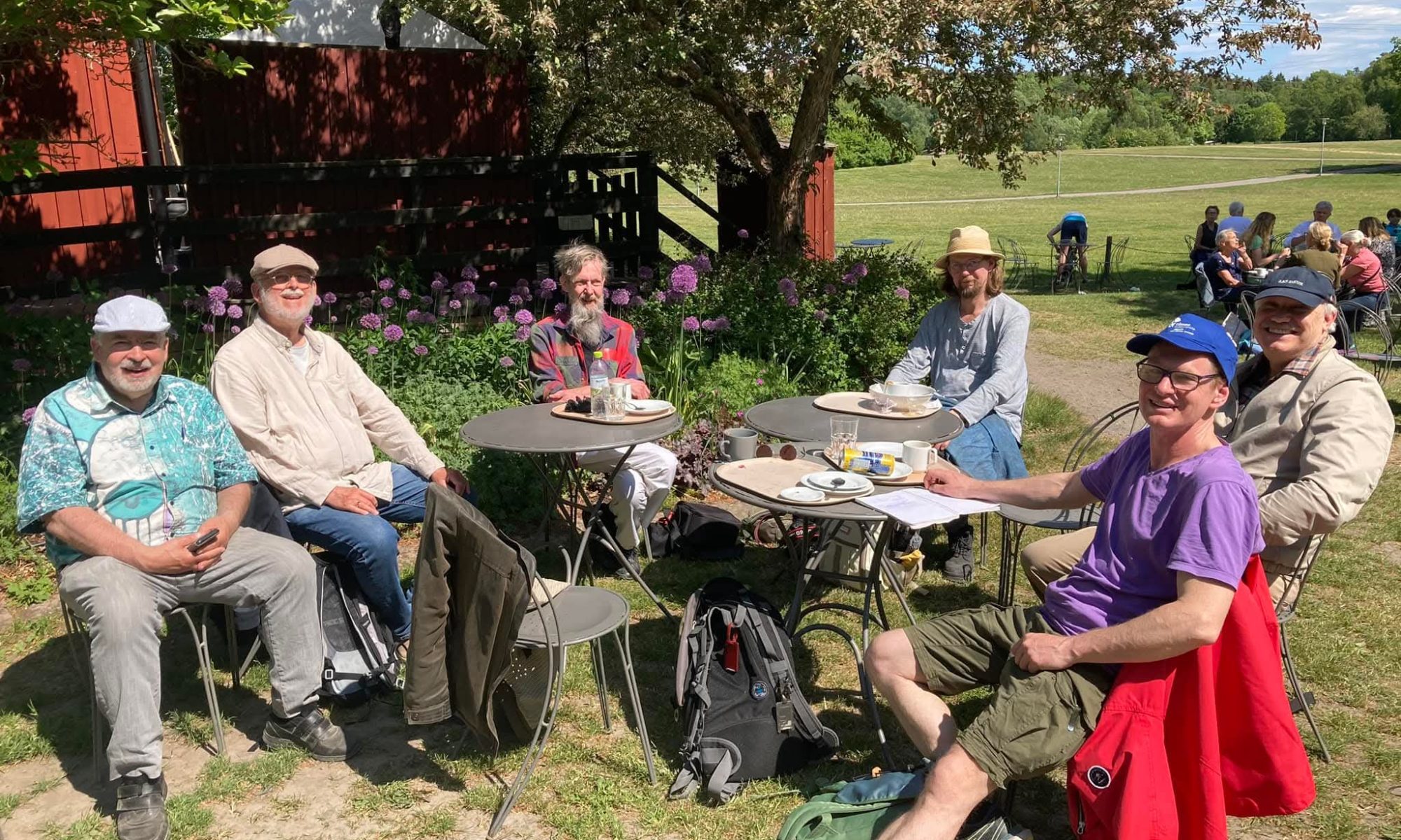 Sex tisdagspoeter sitter kring cafébord i en grönskande trädgård.